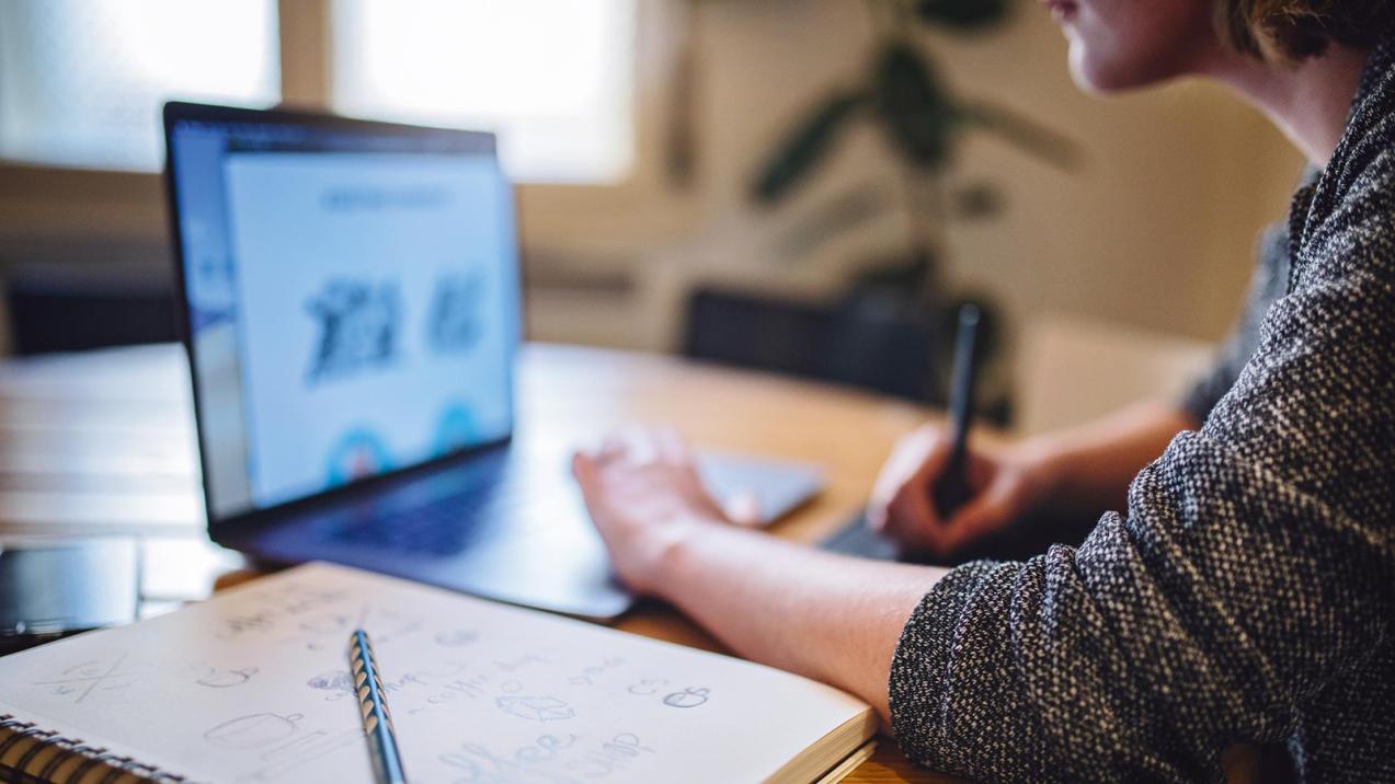 Eine Studentin sitzt an ihrem Laptop am Schreibtisch. Links neben ihr liegt ein Notizblock mit einem Stift. Sie hat auch einen Stift in ihrer rechten Hand und schreibt auf einem Zettel.